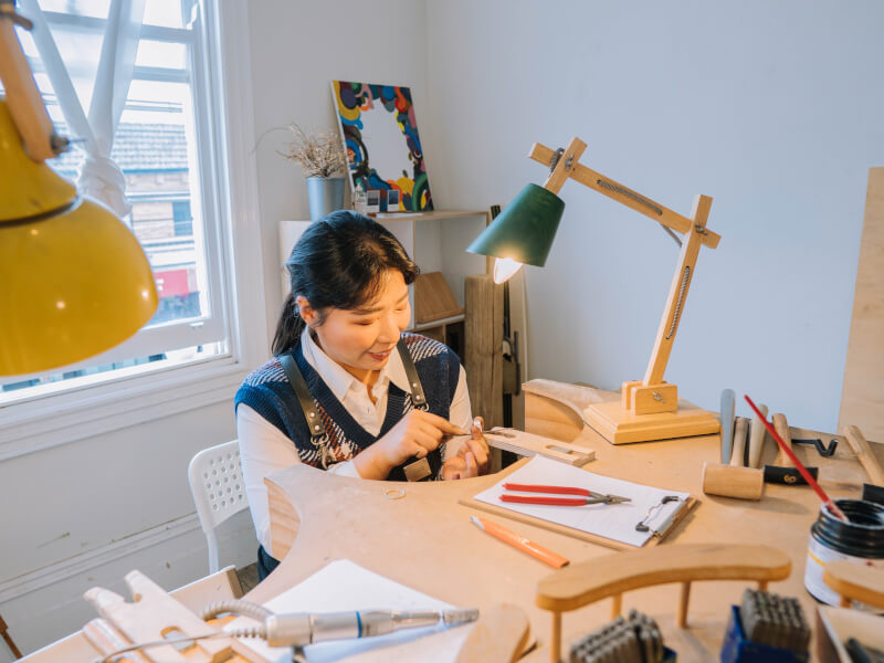 A woman smiles as she looks down at a jewellery project she is working on with a lamp overhead